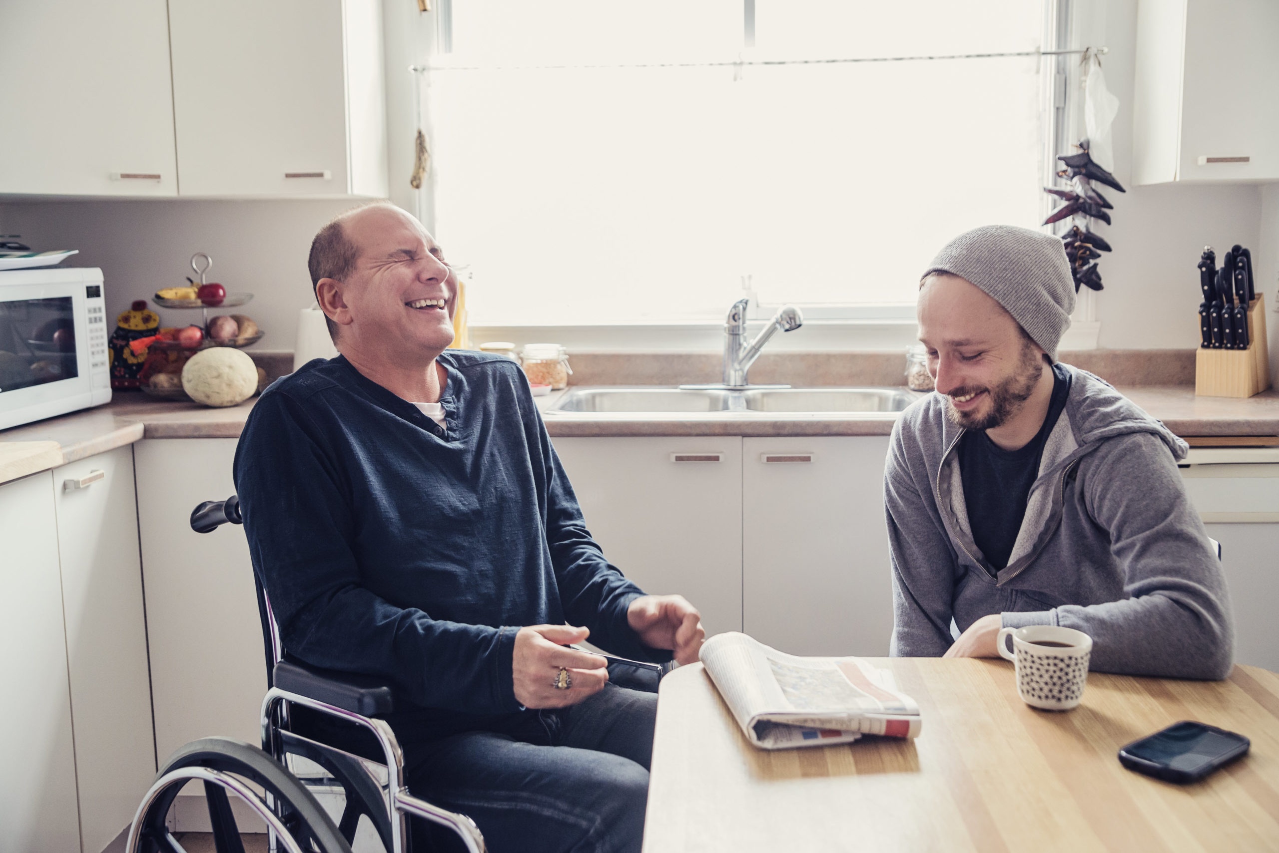 older man seated in wheelcheer laughs with younger man in the kitchen