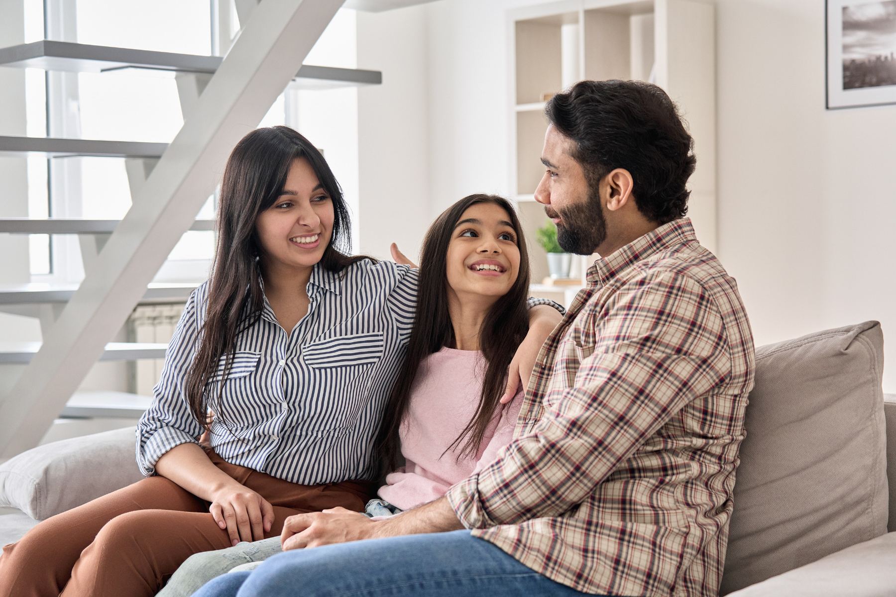 man and woman, sitting on the couch at home with their arms around a young tween girl, in conversation together