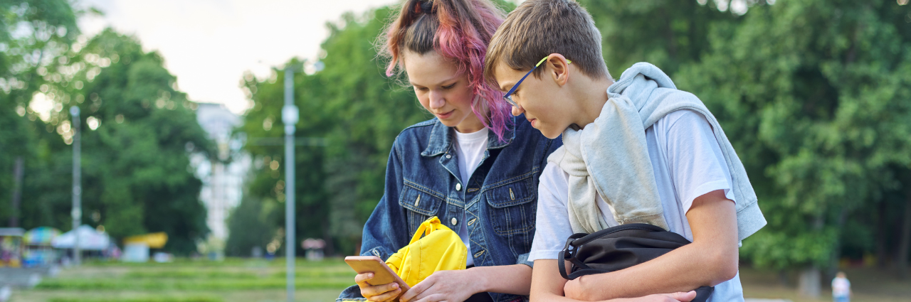 Two teens sitting next to each other outside looking at a phone