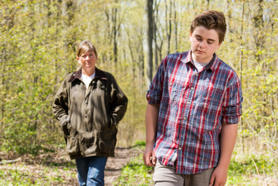 white teen boy, with a plaid shirt and eyes downcast, walks ahead of his mother on a woodland trail