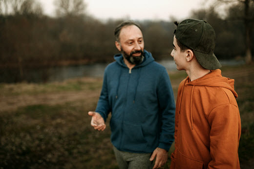 middle-aged father and teen son, wearing hooded sweatshirts, walk by the river in conversation