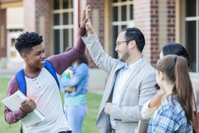 male teacher, wearing glasses and a blazer, high fives with a Black teen student, wearing a henley shirt and a backpack, outside a school building, as other students look on
