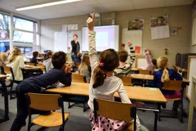 elementary school kids in a classroom raise their hands to be called upon by the teacher