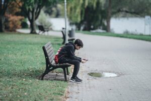 young teen sits alone on a park bench, scrolling on his phone