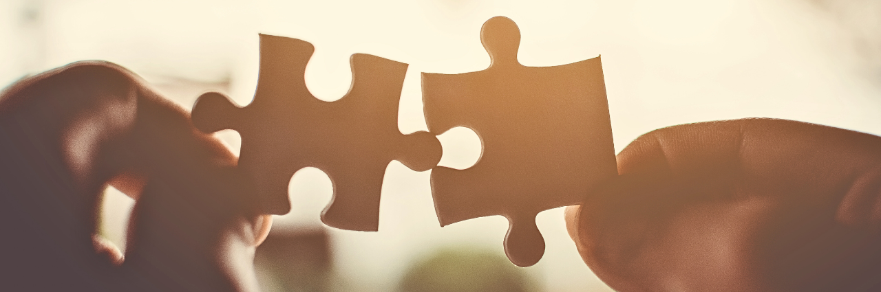 Two hands holding interlocking puzzle pieces, symbolizing connection, teamwork, and problem-solving, with warm sunlight in the background.